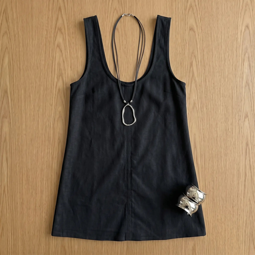 A classic black sleeveless A-line mini dress styled with a silver pendant necklace and cuff bracelets, laid flat on a wooden surface.