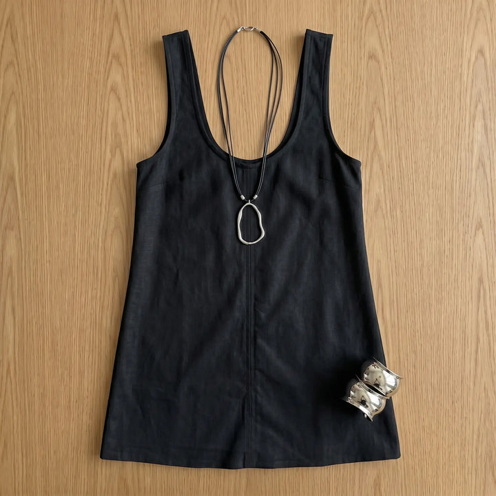A classic black sleeveless A-line mini dress styled with a silver pendant necklace and cuff bracelets, laid flat on a wooden surface.
