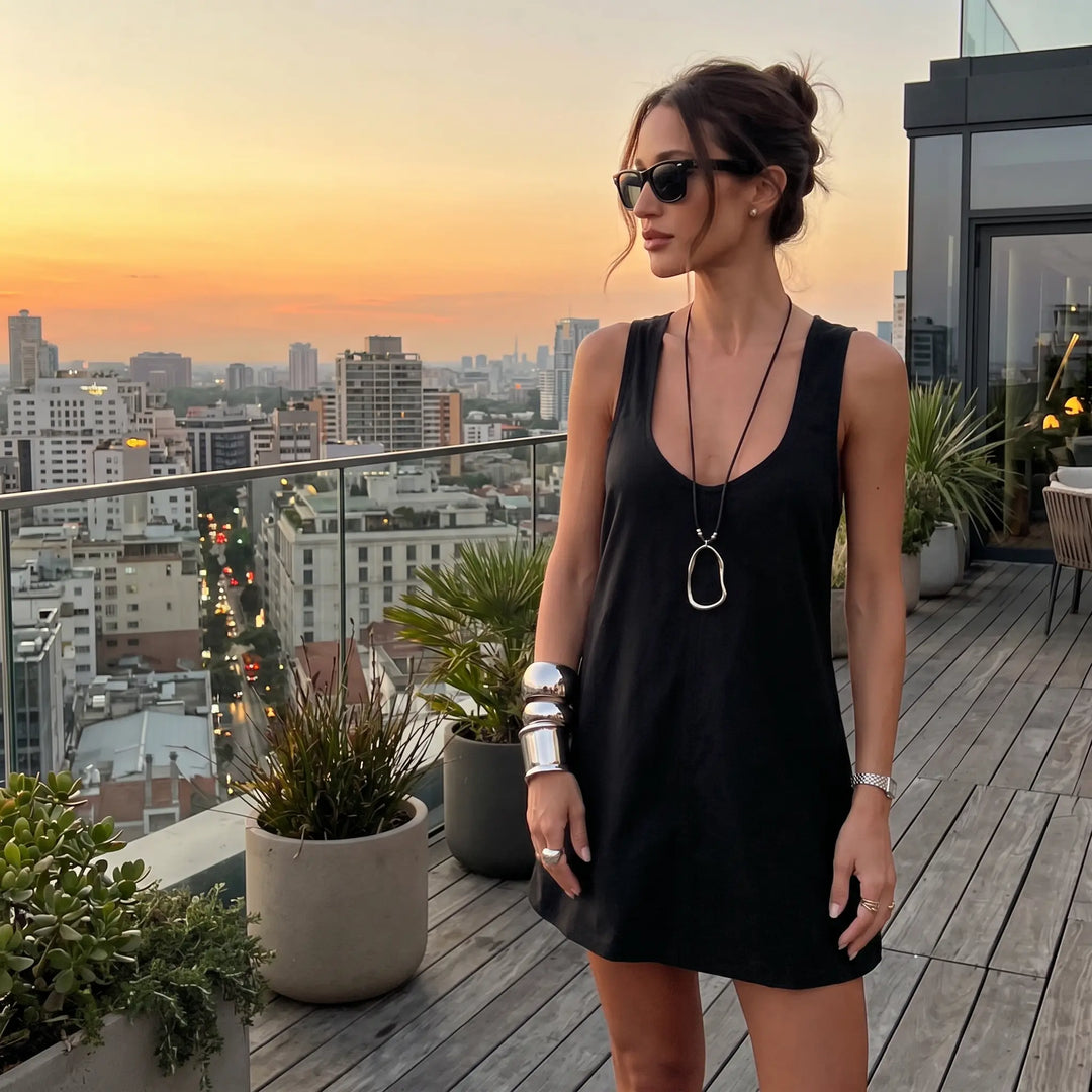 A woman wearing a black sleeveless U-neck A-line mini dress stands on a rooftop at dusk, looking out at a city skyline. The dress is styled with sunglasses, bold jewelry, and a long pendant necklace.
