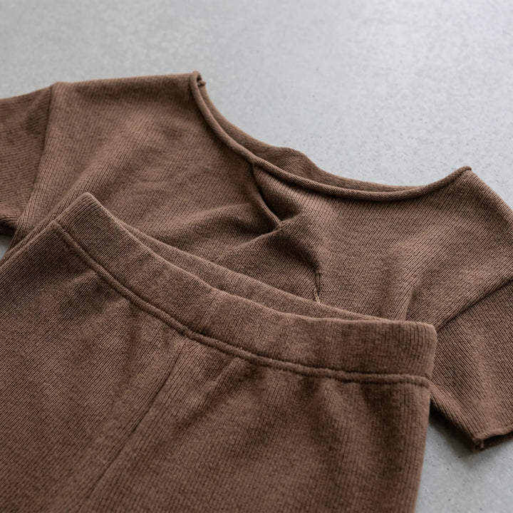 Close-up of a warm deep brown knit loungewear set showcasing fine rib texture and raw frayed edges on the asymmetrical neckline and sleeves.