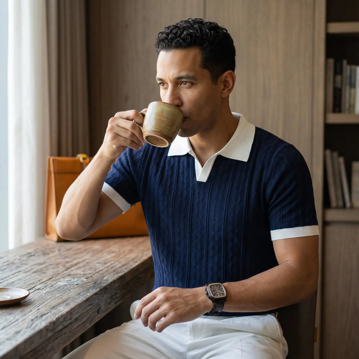 Man wearing a dark blue short-sleeve knitted polo shirt with white collar and cuffs, enjoying coffee at a rustic wooden table in a lifestyle setting.