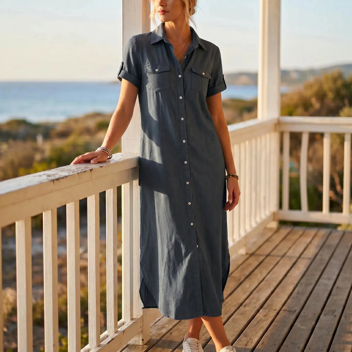A woman models a timeless midi-length, collared, short-sleeved denim shirt dress in a deep charcoal wash, standing on a wooden porch with a coastal ocean view.