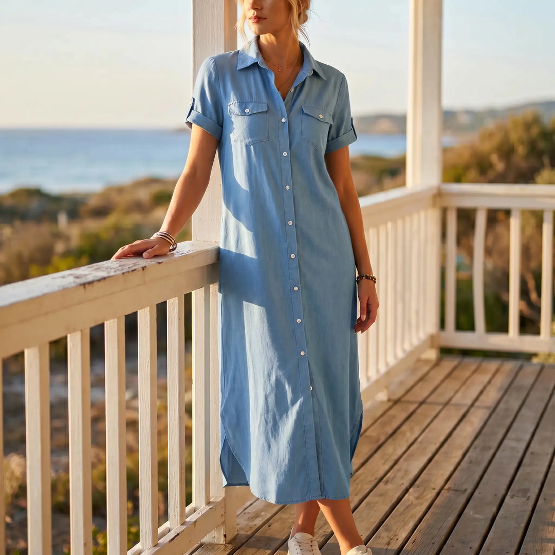 Woman wearing a medium blue wash denim shirt dress with short rolled sleeves and a midi length, standing on a wooden balcony overlooking the ocean.