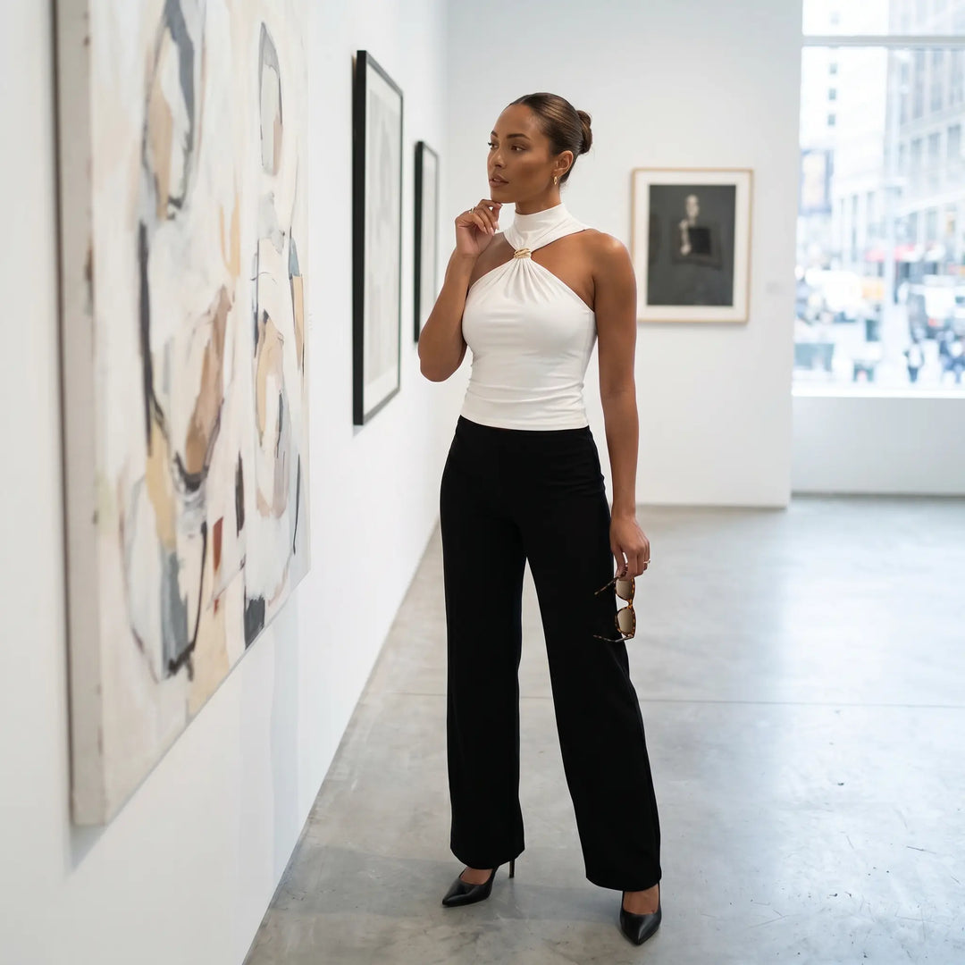 A sophisticated woman in a white halter-neck top with a gold ring detail and black wide-leg pants stands in an art gallery, contemplating a painting. This lifestyle shot captures an elegant, curated moment.