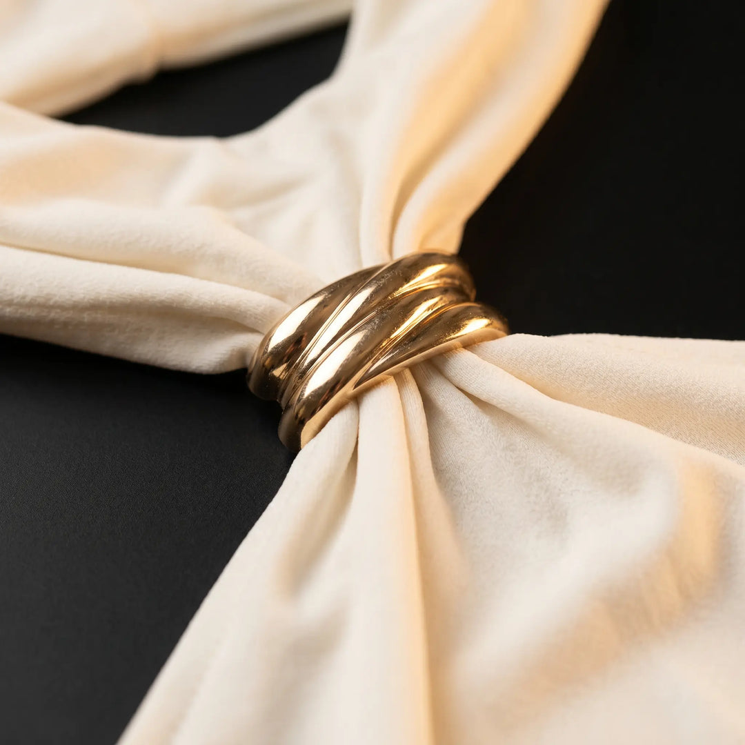 Close-up view of an elegant ivory halter neck top, highlighting the intricate twisted gold-tone metal ring detail and the smooth, slinky stretch-knit fabric with subtle sheen. The fabric drapes artfully around the hardware.