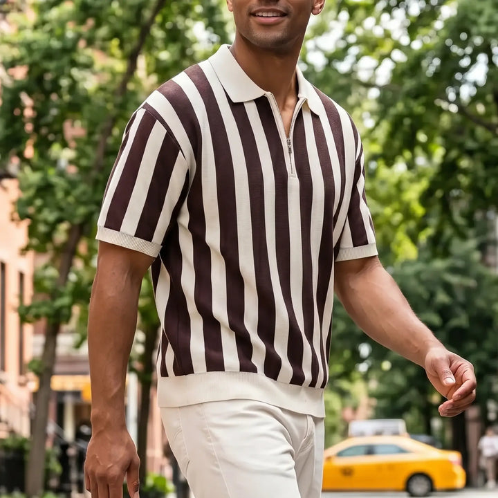 Man walking outdoors wearing a brown and cream vertical striped short-sleeve knit polo shirt with a quarter-zip neckline and light-colored pants, set against a city street background with trees and a yellow taxi.