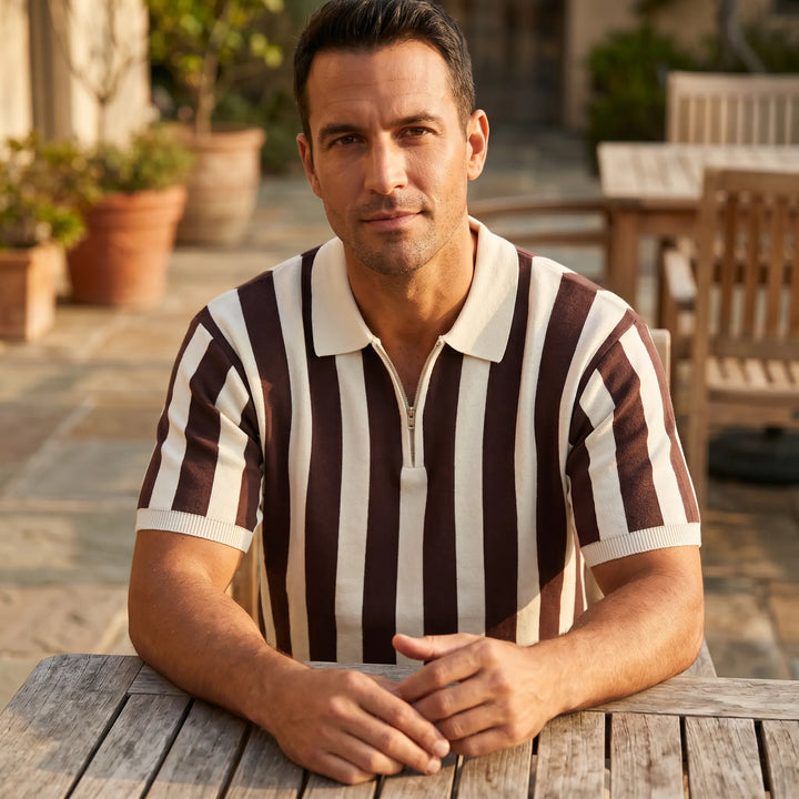 Man in brown and cream vertical stripe knit polo shirt with quarter-zip, seated at a wooden table in an outdoor setting with warm lighting. A classic casual look.