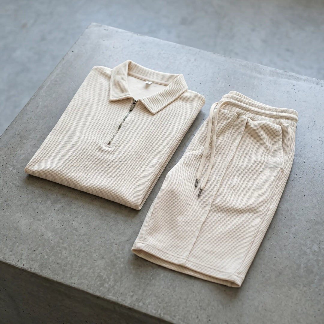 Light beige waffle knit loungewear set featuring a quarter-zip polo shirt and drawstring shorts, presented flat on a textured grey studio surface.
