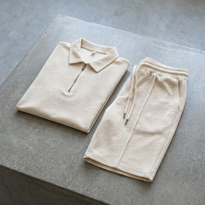 Light beige waffle knit loungewear set featuring a quarter-zip polo shirt and drawstring shorts, presented flat on a textured grey studio surface.