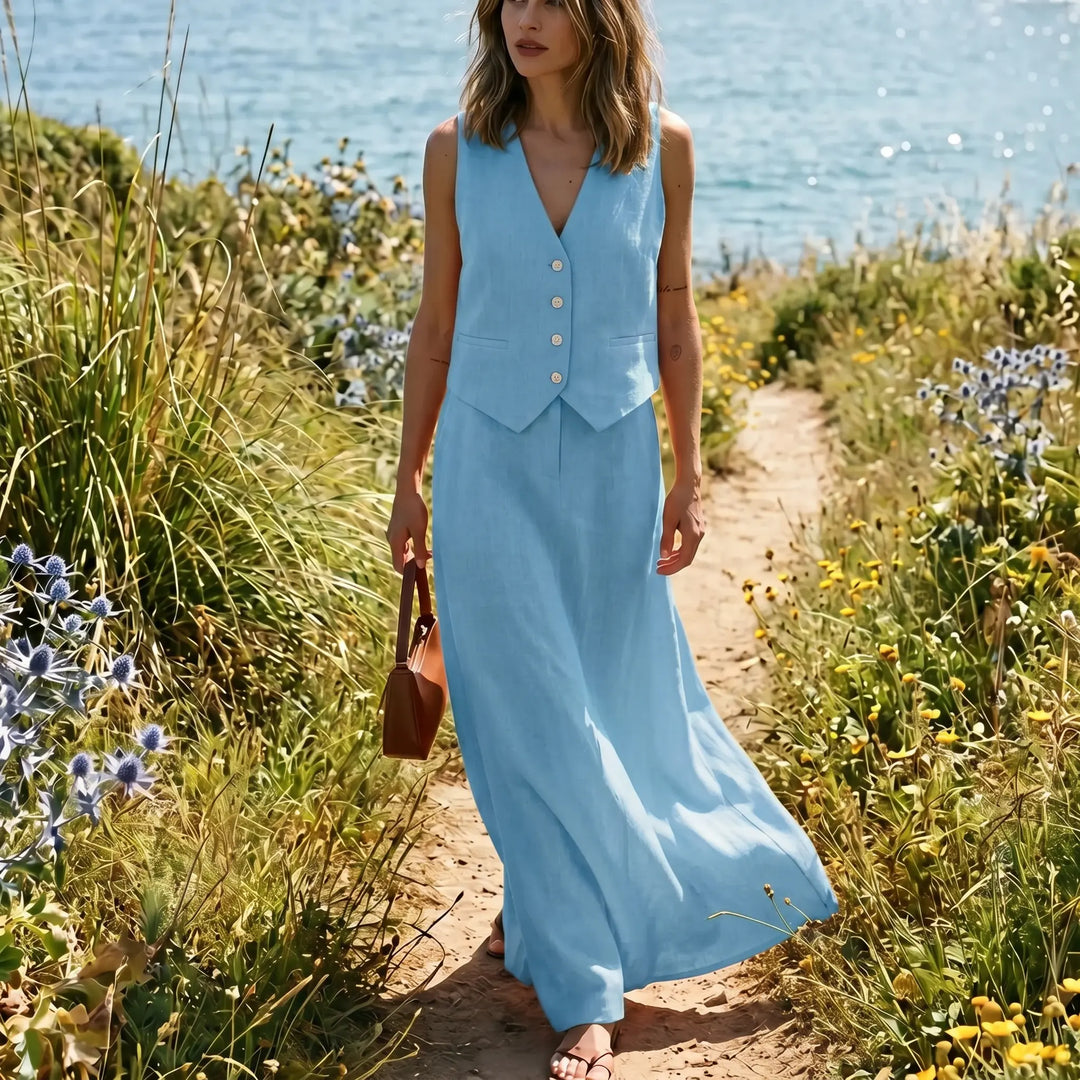 Woman wearing a light blue linen blend sleeveless V-neck vest and a flowing high-waisted maxi skirt, walking on a coastal path with lush greenery and the sea in the background.