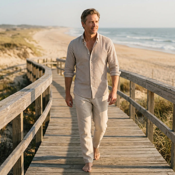 Man wearing a striped linen-blend shirt and light trousers walks on a wooden boardwalk by the beach.