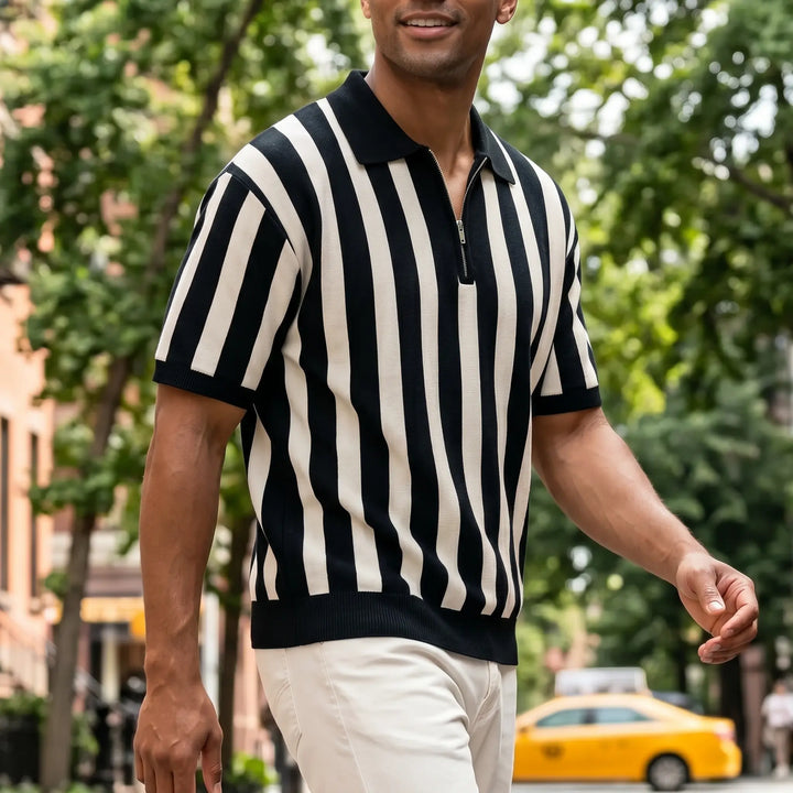 Man wearing a black and cream vertical striped knit polo shirt with a quarter-zip neckline and a relaxed fit, walking outdoors in a city.