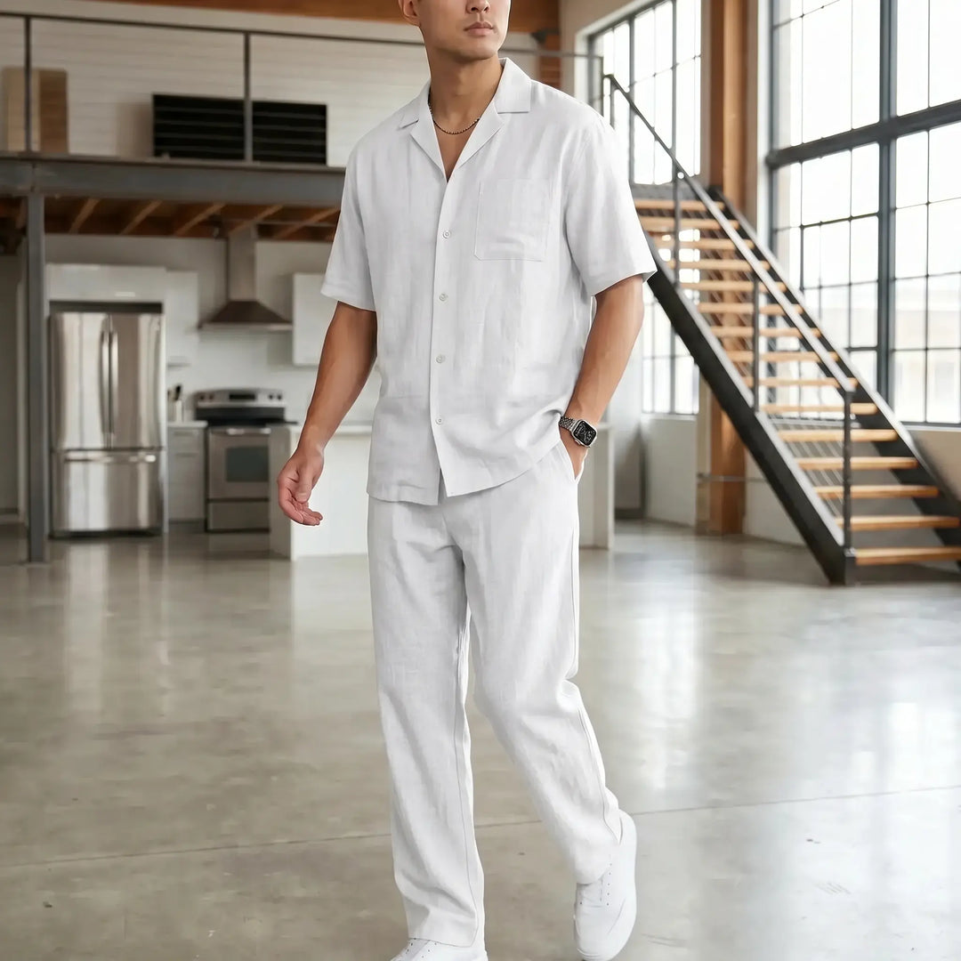 Men's crisp white linen blend casual set featuring a camp collar shirt and straight-leg trousers, modeled in a contemporary interior lifestyle setting with modern kitchen and stairs.