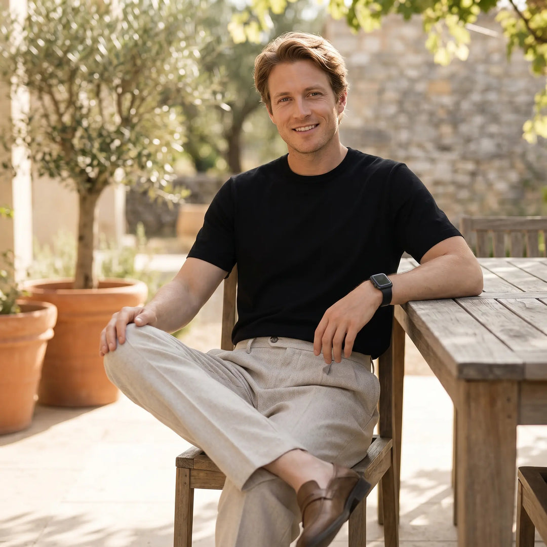 A man with fair skin and blond hair smiles while sitting outdoors, wearing oat beige Mirok men&