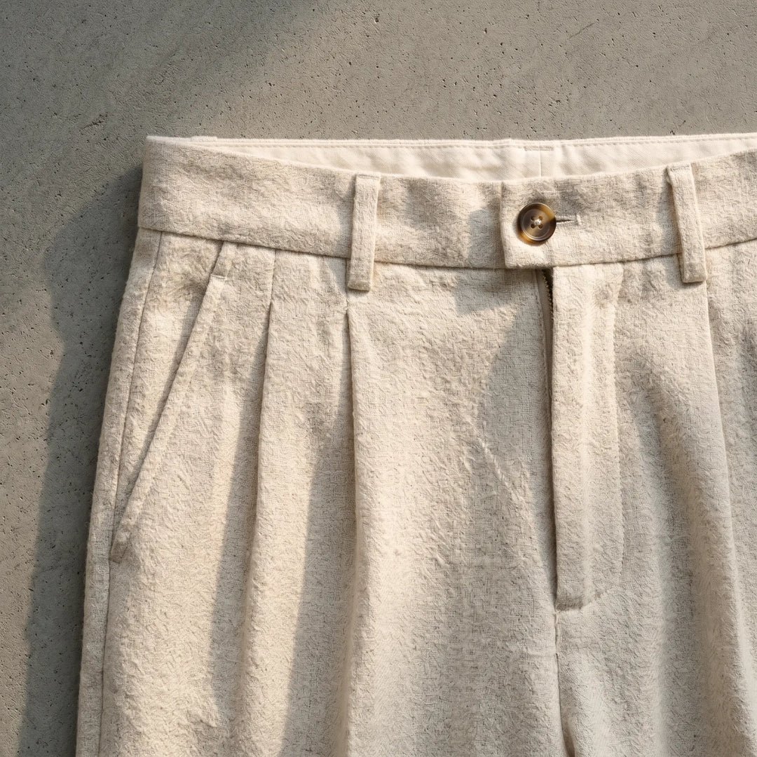 Close-up of oat-beige wool-blend trousers featuring a subtle herringbone weave, single front pleats, button-tab closure, and diagonal front pockets.