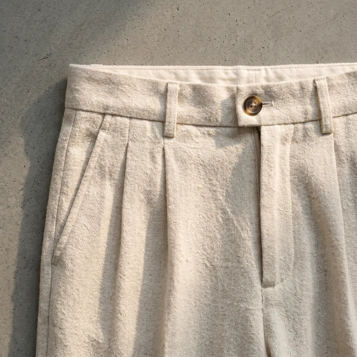 Close-up of oat-beige wool-blend trousers featuring a subtle herringbone weave, single front pleats, button-tab closure, and diagonal front pockets.