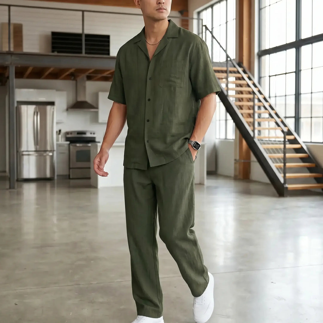 A man wearing an olive green linen-blend casual set, featuring a camp collar shirt and straight-leg trousers, styled in a loft interior. He is looking away from the camera with his hands in his pockets.