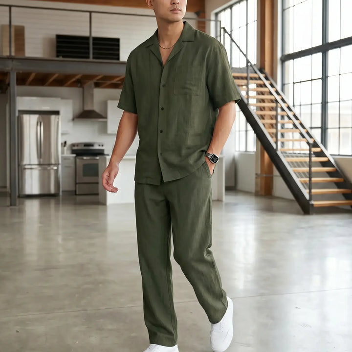 A man wearing an olive green linen-blend casual set, featuring a camp collar shirt and straight-leg trousers, styled in a loft interior. He is looking away from the camera with his hands in his pockets.