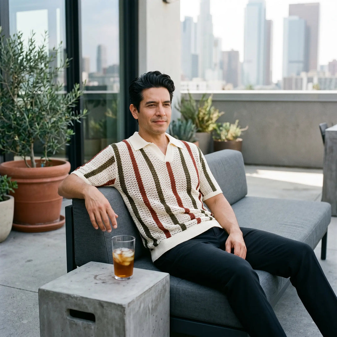 A man relaxing on a modern loft terrace wears a Rysol short-sleeved knit polo shirt featuring bold vertical stripes in cream, rust, and olive green, paired with black trousers.