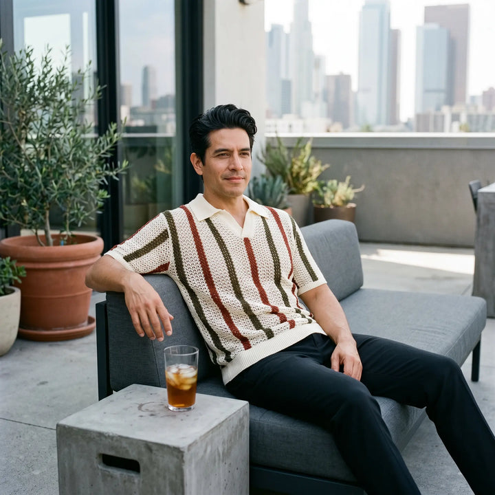 A man relaxing on a modern loft terrace wears a Rysol short-sleeved knit polo shirt featuring bold vertical stripes in cream, rust, and olive green, paired with black trousers.
