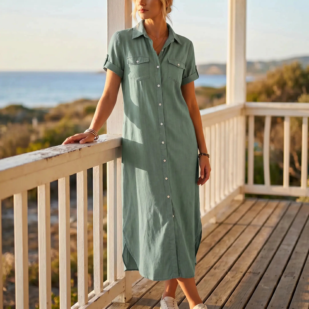 A woman wears a sage green midi denim shirt dress with short rolled sleeves and side slits, standing on a wooden porch with a coastal view.