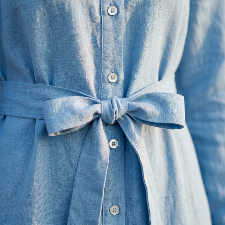 Close-up of a bright sky blue linen-blend shirt dress, showcasing the subtle vertical rib texture, pearlized white button-front closure, and a tied fabric self-tie belt.