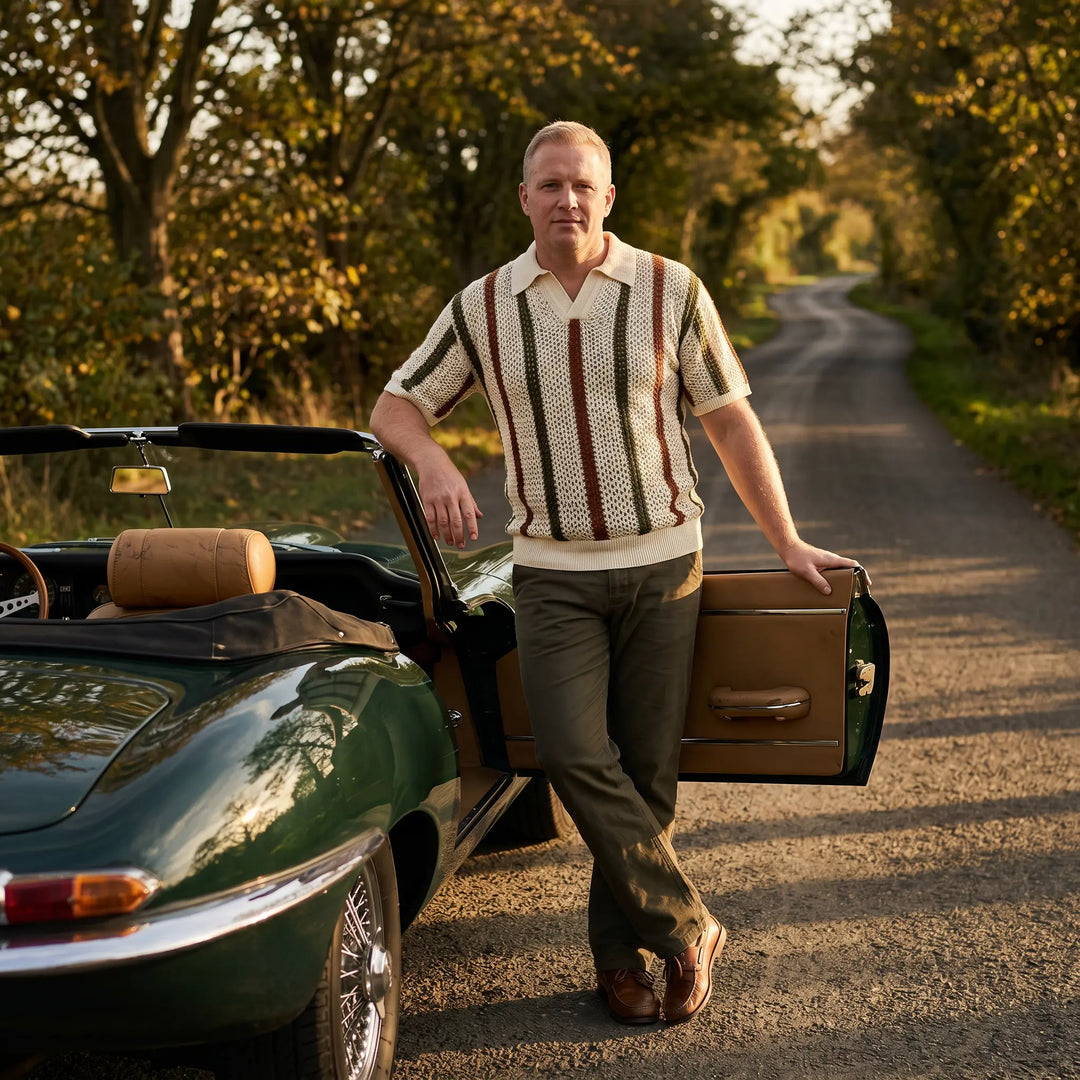 Man wearing a cream, rust, and dark green striped open-knit polo shirt with a V-neck and ribbed collar, leaning on a classic convertible car on a scenic road.