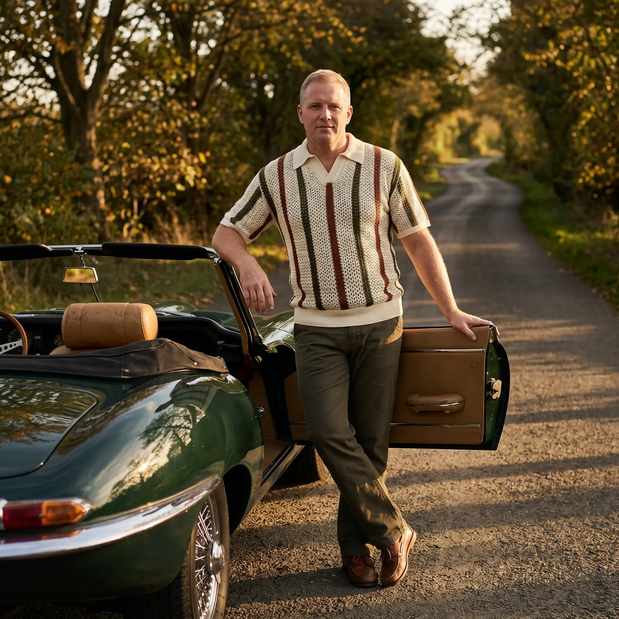 Man wearing a cream, rust, and dark green striped open-knit polo shirt with a V-neck and ribbed collar, leaning on a classic convertible car on a scenic road.