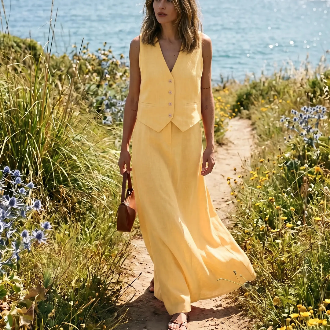 Woman wearing a vibrant sunshine yellow linen-blend vest and flowing maxi skirt set, walking on a path by the sea. The cropped vest features a V-neck and natural buttons, while the high-waisted skirt drapes elegantly.