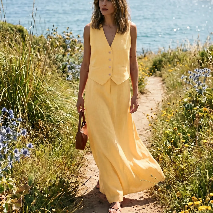 Woman wearing a vibrant sunshine yellow linen-blend vest and flowing maxi skirt set, walking on a path by the sea. The cropped vest features a V-neck and natural buttons, while the high-waisted skirt drapes elegantly.