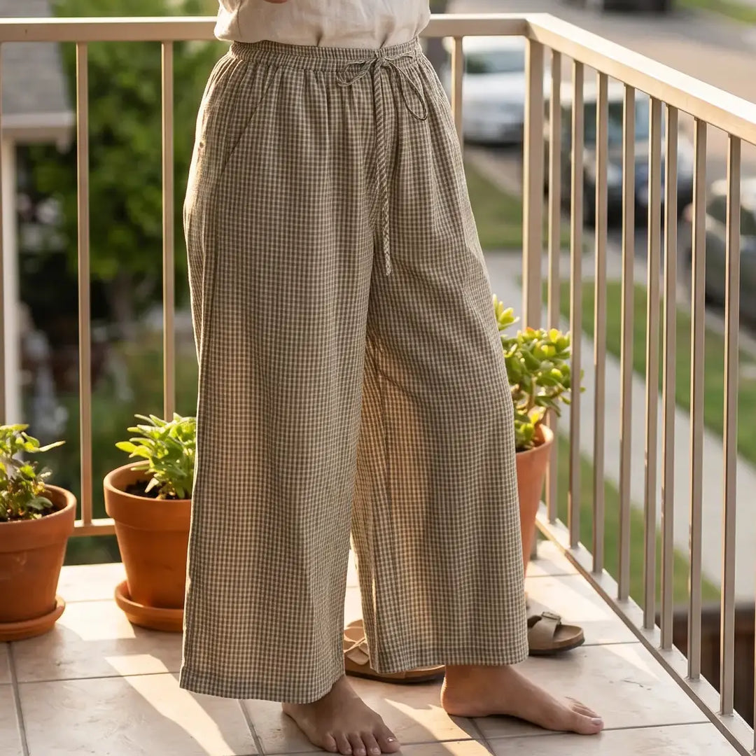 Woman wearing Zelphyr wide-leg plaid trousers and a tank top, enjoying a drink on a sunny balcony with a suburban street view.