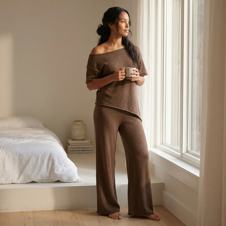A woman in a deep brown ribbed knit loungewear set with an asymmetrical off-shoulder top and wide-leg pants holds a mug by a sunlit window in a serene bedroom.