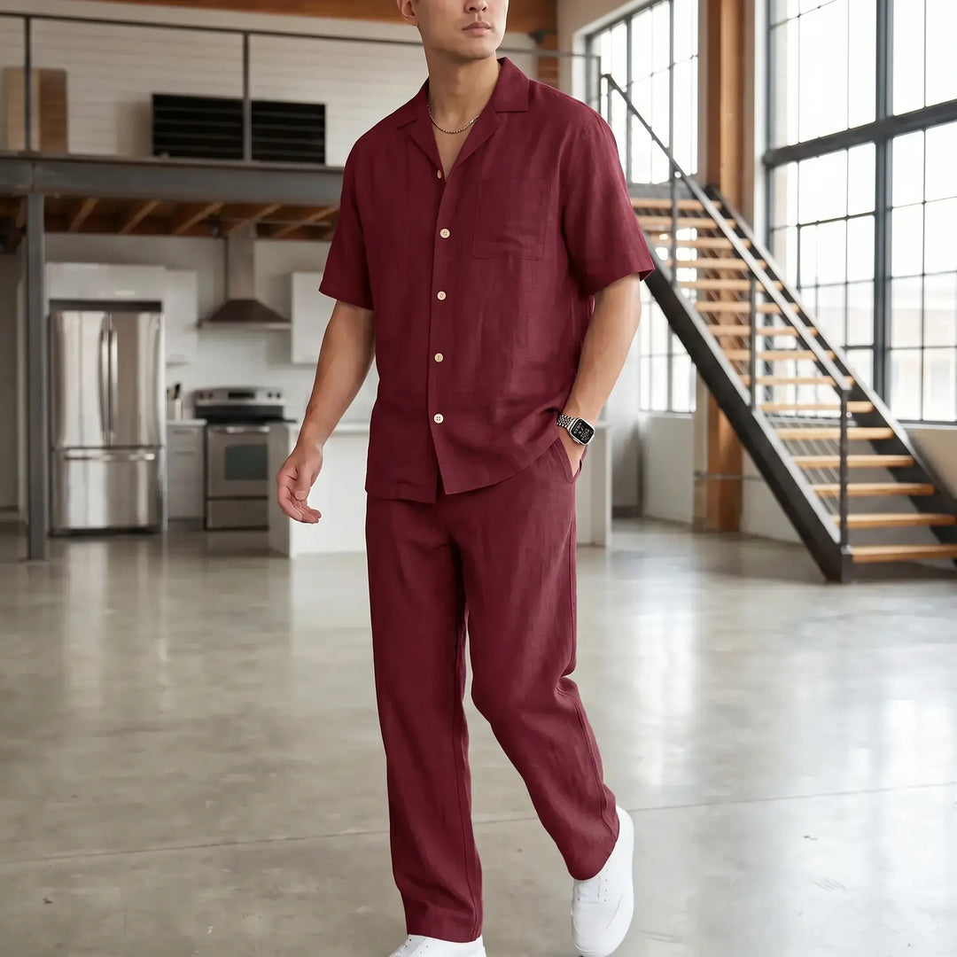 A man models a rich burgundy linen-blend casual set, featuring a short-sleeve camp collar shirt and straight-leg trousers, standing in a modern loft interior with hands in pockets.
