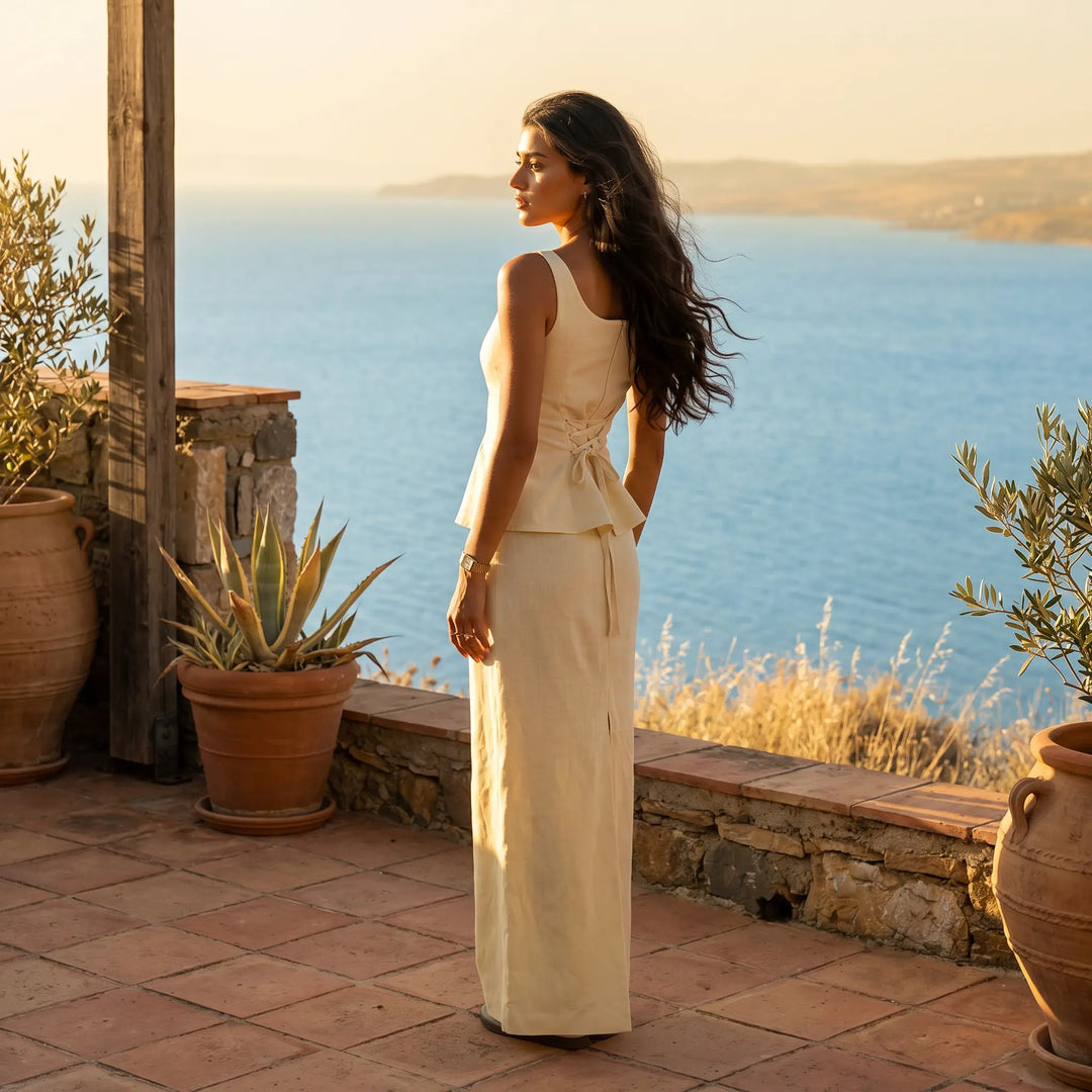 Woman wearing a pale yellow linen-blend peplum top and maxi skirt set, standing on a terrace overlooking the sea at sunset.
