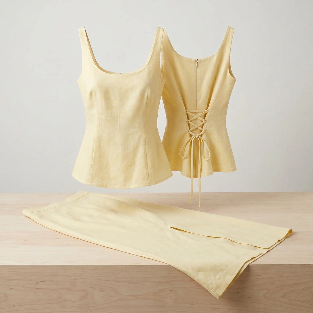 Pale warm yellow linen-blend sleeveless peplum top with a scoop neckline and lace-up back, paired with a matching high-waisted maxi skirt featuring a side slit. Styled as a two-piece set on a studio background.