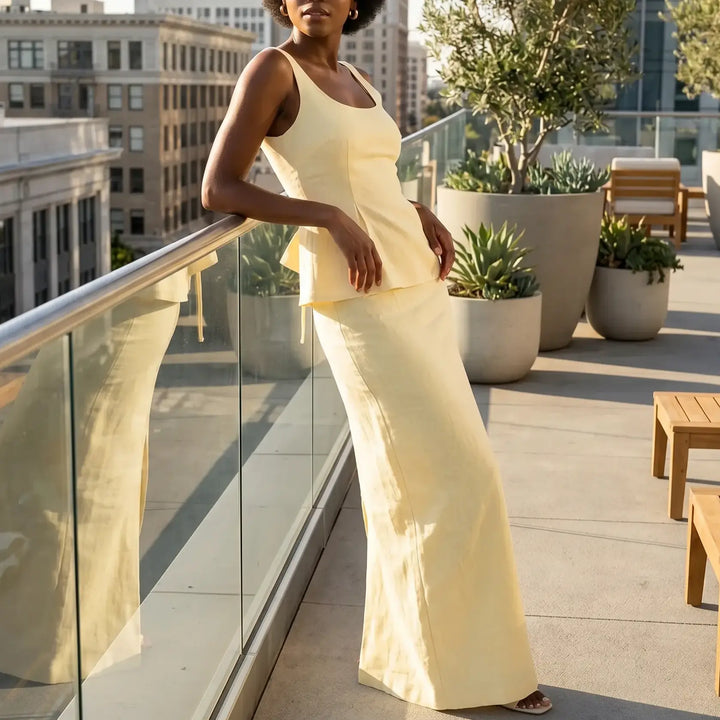 Woman wearing a pale yellow linen-blend peplum top and maxi skirt set, posing on a city rooftop with a serene expression.