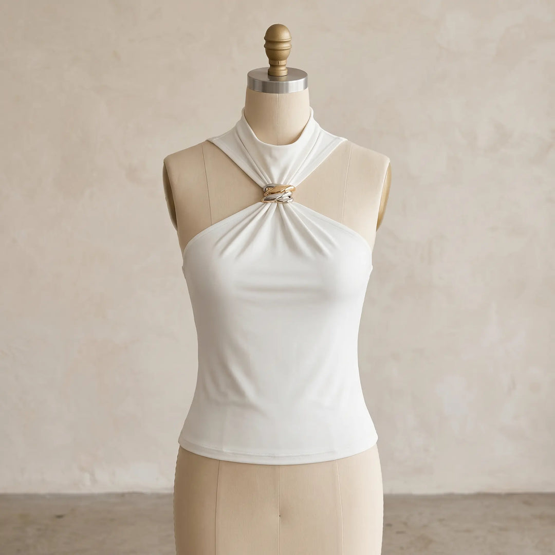 Creamy ivory sleeveless halter neck top featuring a twisted gold-tone metal ring detail at the neckline, presented on a mannequin with a textured studio background. This elegant knit blouse offers a body-skimming fit.