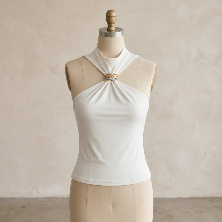 Creamy ivory sleeveless halter neck top featuring a twisted gold-tone metal ring detail at the neckline, presented on a mannequin with a textured studio background. This elegant knit blouse offers a body-skimming fit.